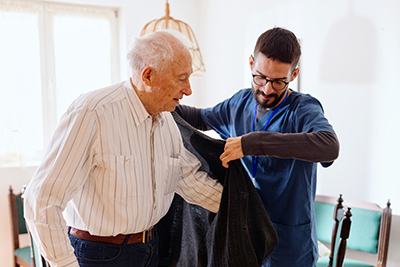 A male caregiver is helping a male senior into his jacket in his home.