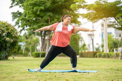 An overweight woman in a park doing yoga on a mat.