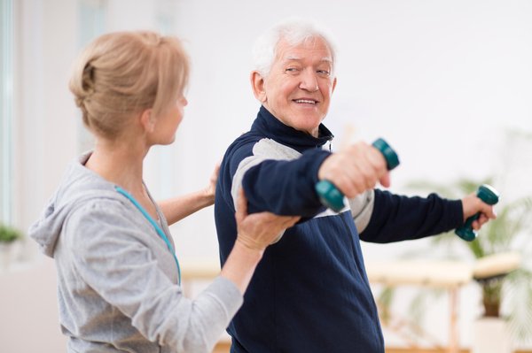 Female fitness trainer working out with a male adult senior, who is lifting dumbbells
