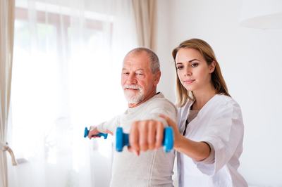 A senior white male exercising with light dumbbells while being assisted by of a female physical therapist.